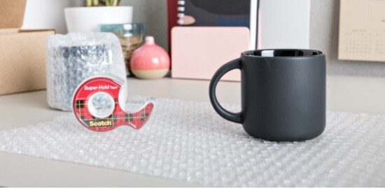 A mug and roll of Scotch® Super-Hold Tape sitting on a sheet of bubble wrap.
