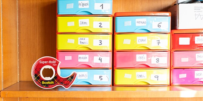 Colorful labeled stationary drawers on a shelf with Scotch® Super-Hold Tape in the foreground.
