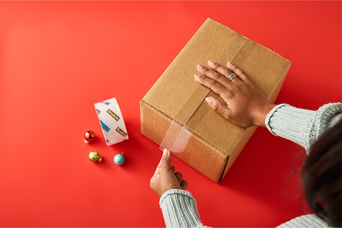 Hands sealing a cardboard box with tape on a red surface, surrounded by small ornaments and a tape dispenser.
