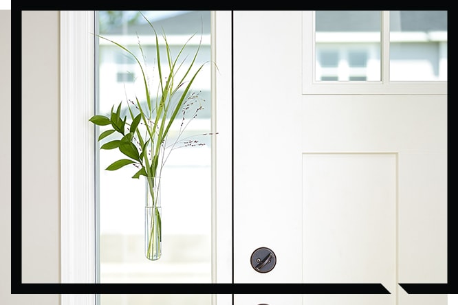 Minimalist white door with a vertical window featuring a glass vase holding green leaves and grass, framed in black.
