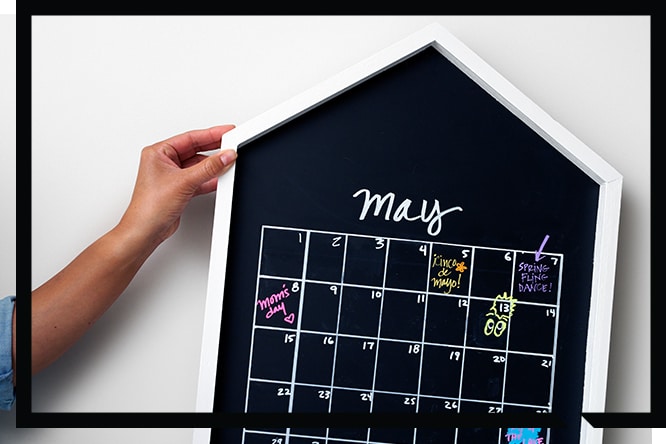 Hands holding a house shaped chalkboard calendar against a wall.

