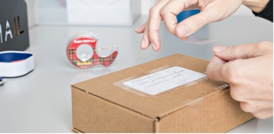 A person securing a mailing label to a cardboard box with Scotch® Super-Hold Tape.
