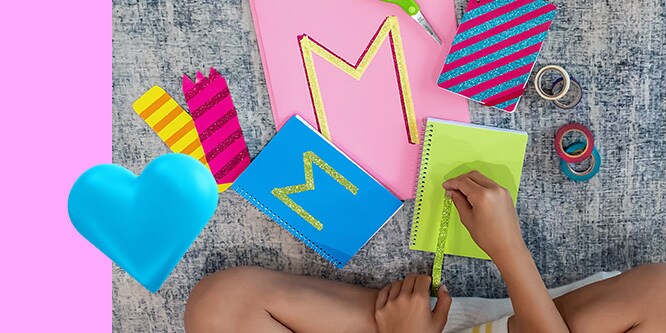 A kid putting Scotch® Fun Tape on  colorful notebooks in the shape of the letter “M”. A blue heart emoji in the lower left corner.
