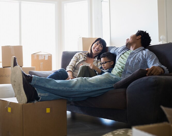 Family relaxing on a sofa in a new home surrounded by moving boxes, with light streaming in through windows.
