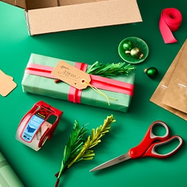 Gift wrapping supplies on a green surface, including a box, wrapped gift, ribbon, scissors, tape, and tags.