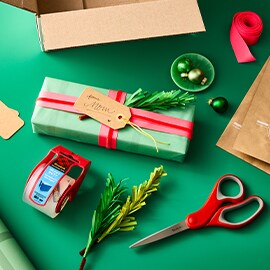 Gift wrapping supplies on a green surface, including a box, wrapped gift, ribbon, scissors, tape, and tags.