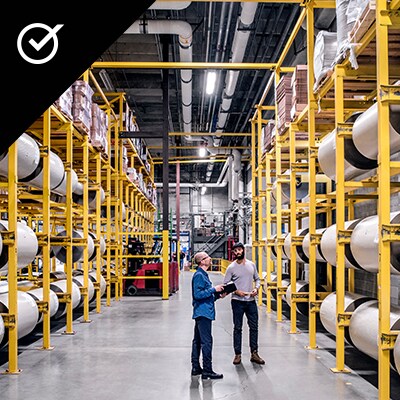 An image of a product manufacturing warehouse, with two people talking. An Icon representing the commitment pillar in the top left: a checkmark in a circle.
