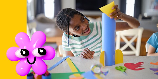 A child creating a paper rocketship at a craft table. A pink 3D flower emoji in the foreground.
