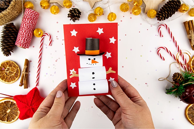 Hands holding a festive card with a snowman design surrounded by Christmas decorations like pinecones and candy canes.
