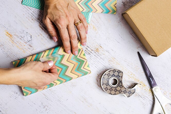 Hands wrap a gift with chevron-patterned paper, surrounded by tape, scissors, and a brown box on a rustic white surface.
