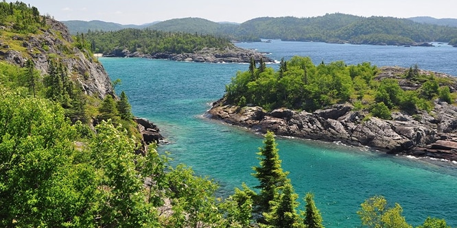 A blue lake landscape from above, green pine trees and cliffs surround the lake.
