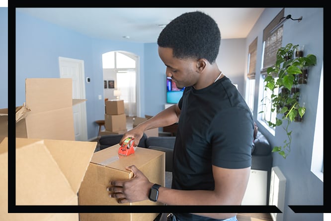 A man surrounded by moving boxes, sealing a cardboard box shut with Scotch® Heavy Duty Moving Packing Tape
