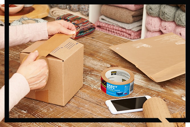 Hands sealing a cardboard box with tape on a wood table, next to Scotch® Heavy Duty Paper Tape.

