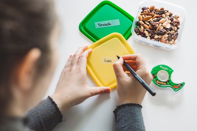 A person labels a yellow lunch container and prepares snacks in a plastic box on a table.

